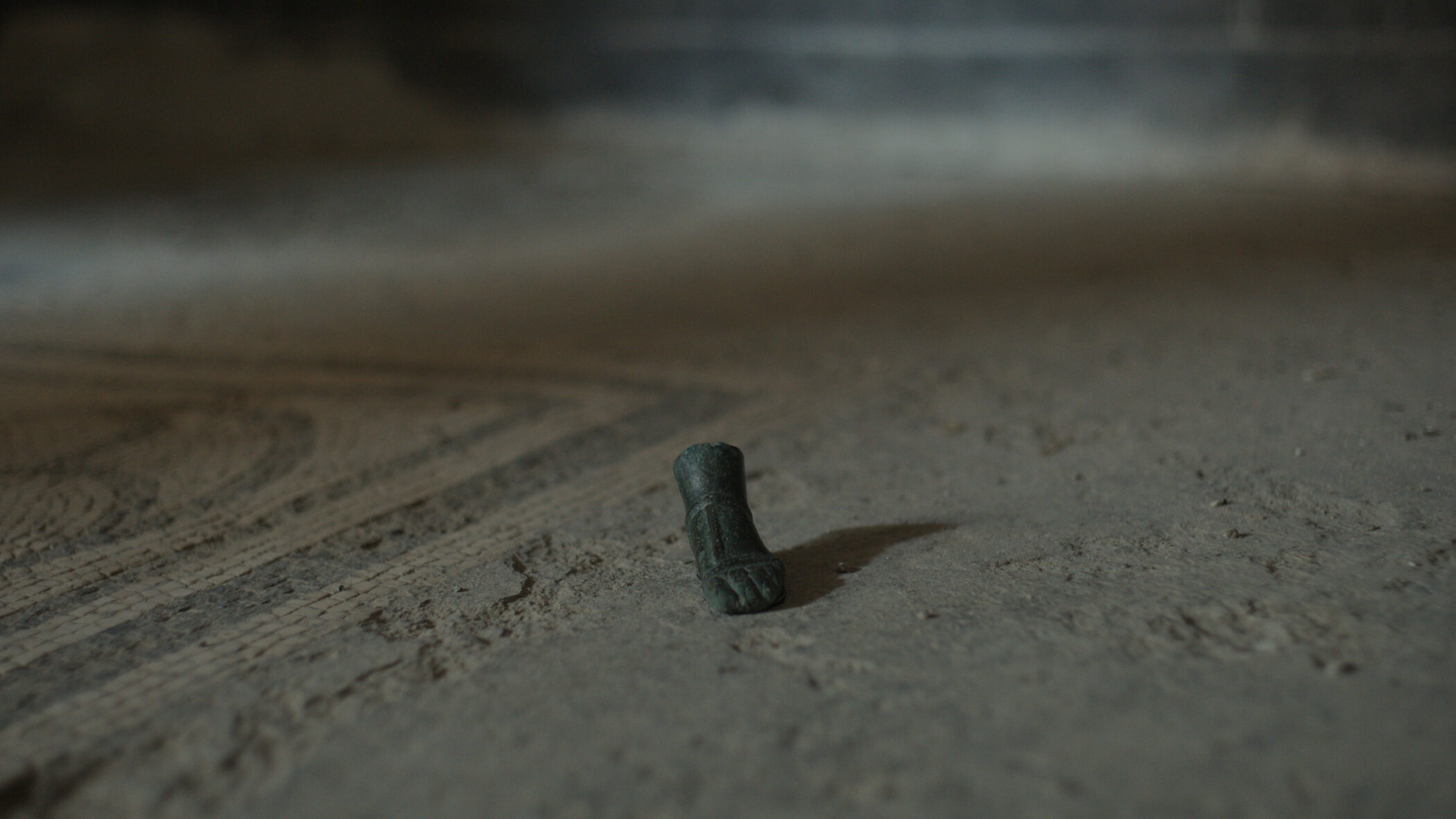 A small bronze foot fragment standing on a dusty ancient mosaic floor in low light