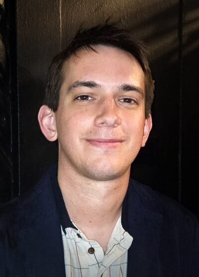 Andrew Dismukes smiling in a navy jacket outside the Upright Citizens Brigade Theatre in New York, March 2025