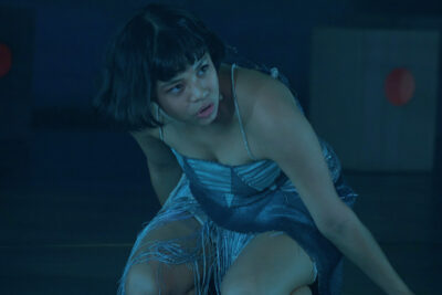 A performer in a fringed blue costume crouches on a dark stage in AYLA A New Musical