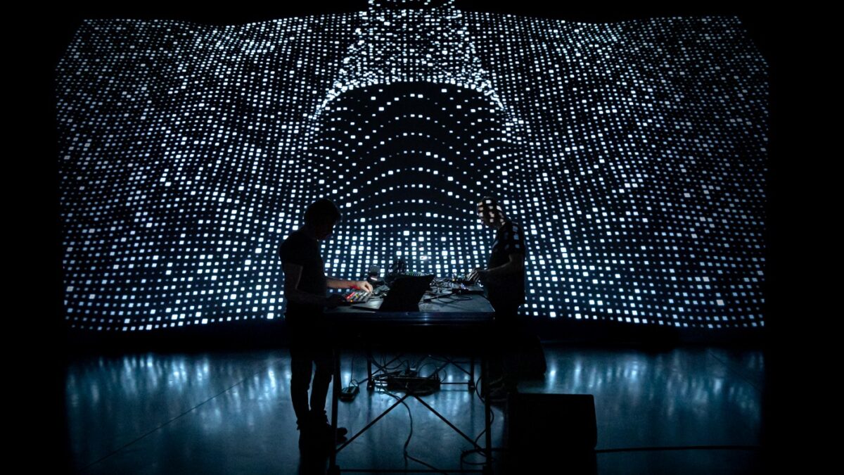 Two performers silhouetted at a table of electronics before a vast screen of white pixel patterns, ATOTAL by Franck Vigroux and Antoine Schmitt