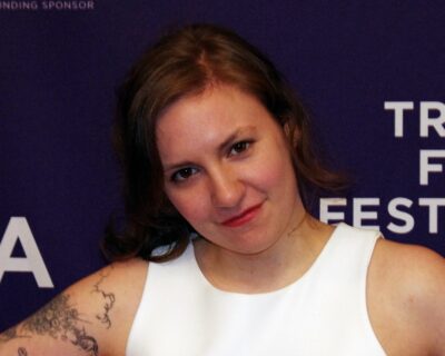 Lena Dunham smiling in a white sleeveless top in front of a Tribeca Film Festival backdrop