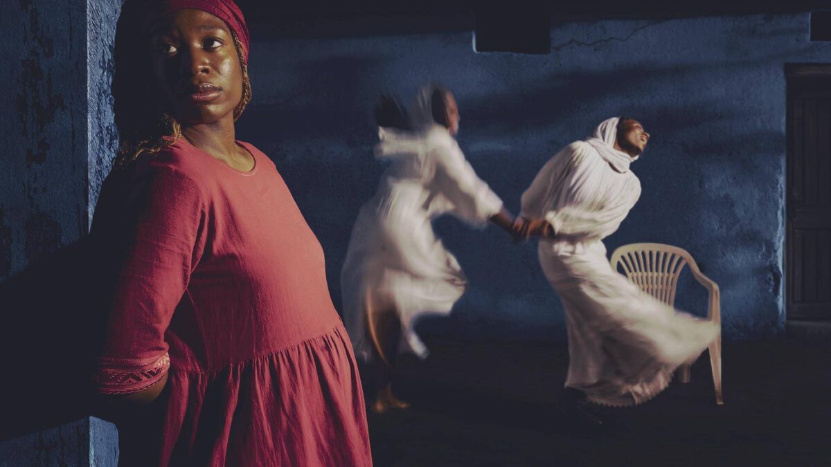 A young woman in a red dress leans against a blue wall while two figures in white robes spin behind her