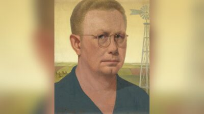 Grant Wood self-portrait in round wire glasses and a dark work shirt, with Iowa fields, haystacks and a windmill behind him