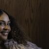 Joy Denalane smiling in a feathered coat against a wood-panelled wall
