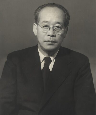 Kenji Mizoguchi in a studio portrait from the 1950s, wearing round glasses and a dark suit