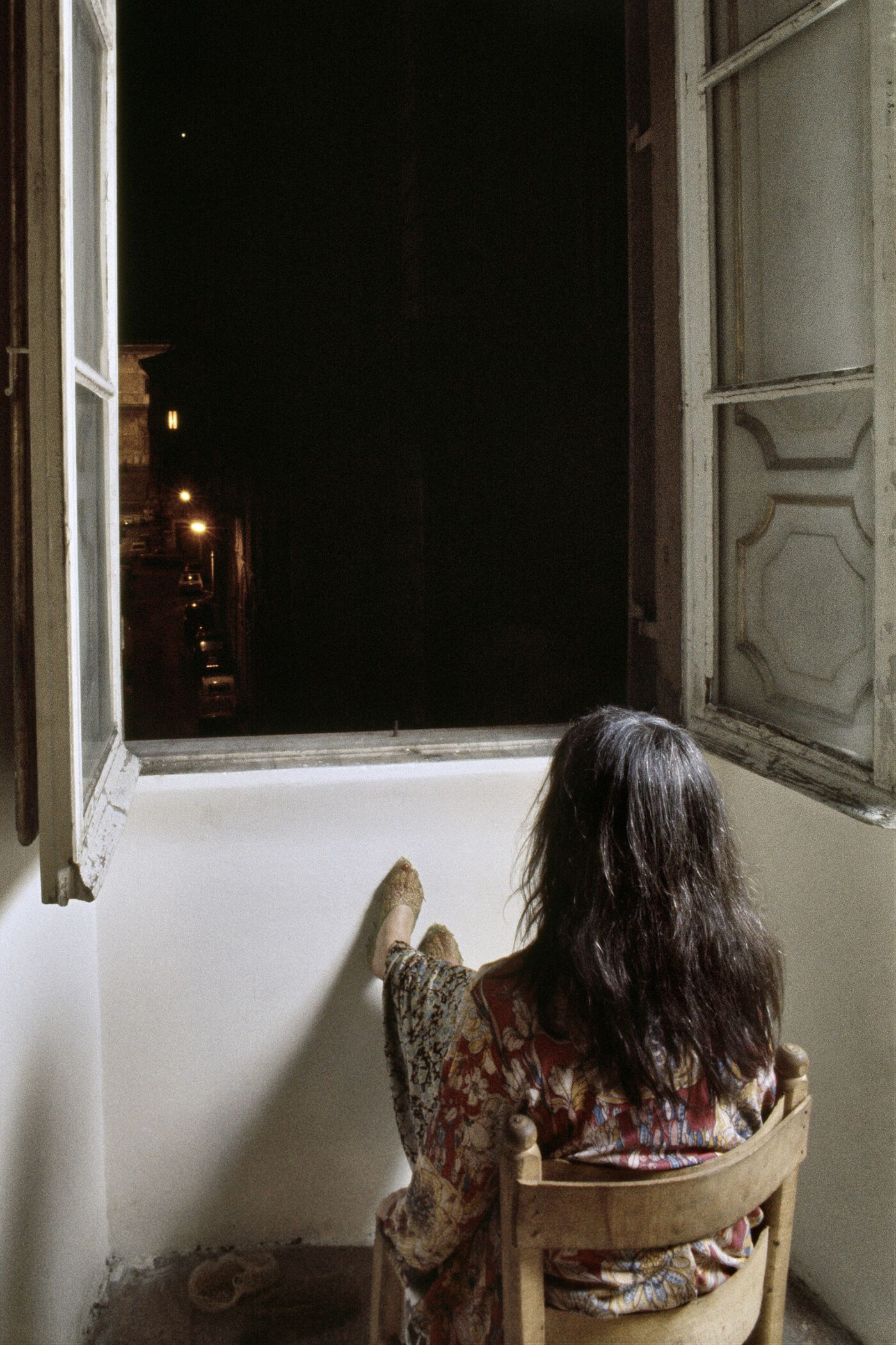 Marisa Merz seen from behind, seated by an open window at night, her feet in knitted slippers raised against the wall