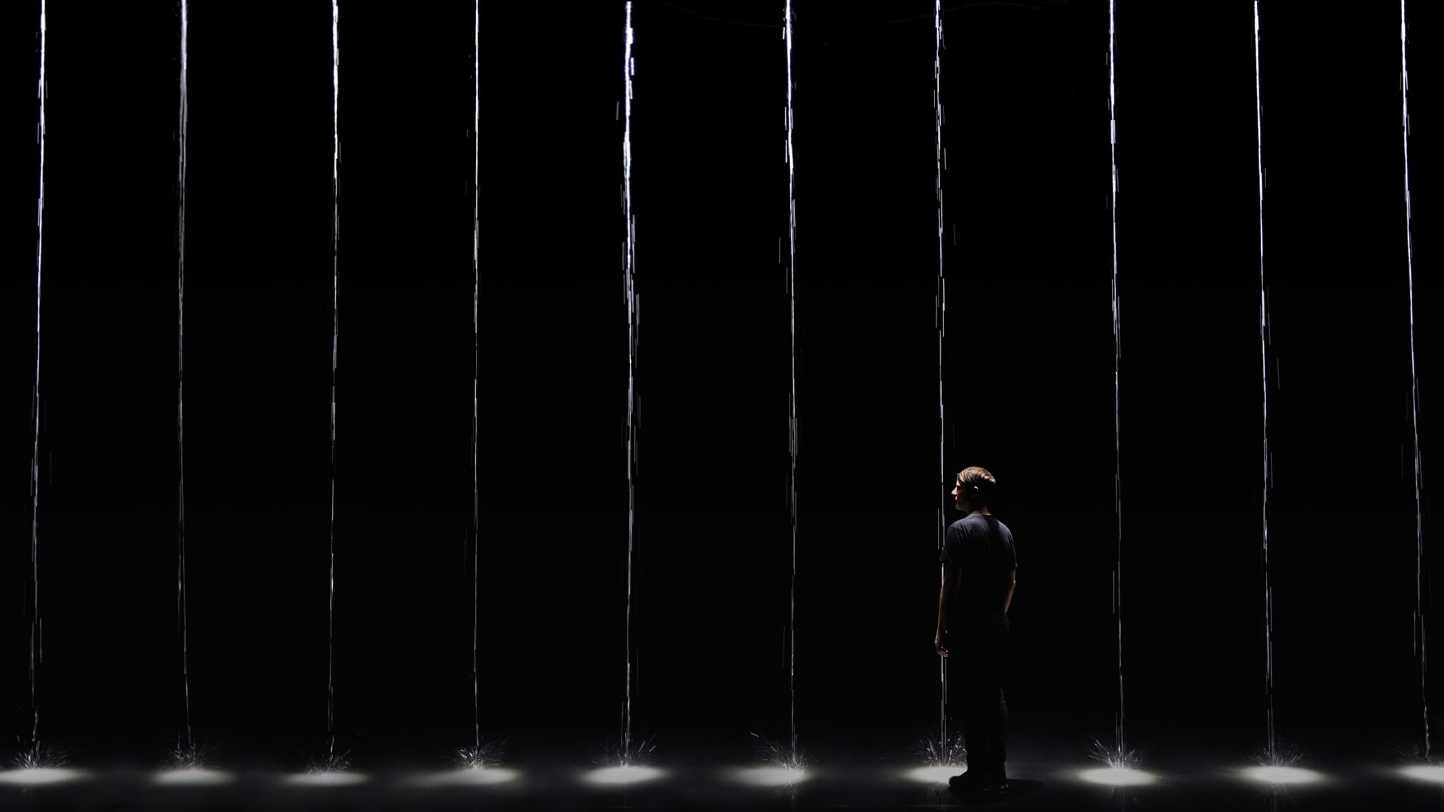 A lone figure stands among thin falling columns of water lit from below in a dark space, Martin Messier installation 1 drop 1000 years