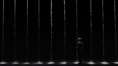 A lone figure stands among thin falling columns of water lit from below in a dark space, Martin Messier installation 1 drop 1000 years