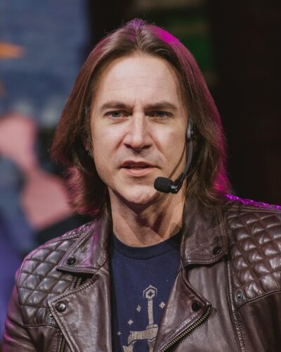 Matt Mercer speaking into a headset microphone on stage at IGN Live 2024, wearing a brown leather jacket