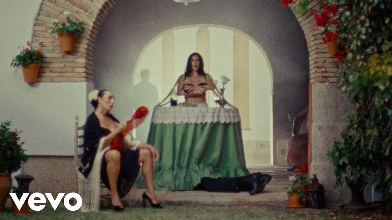 Judeline stands behind a draped table in an Andalusian courtyard while Rossy de Palma sits with a fan in the La Falda video