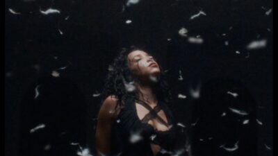 Tinashe in a dark room under falling white feathers in the Pillow Fight music video