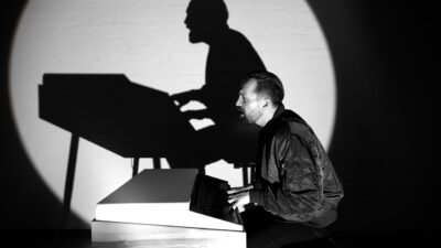 Danger Dan playing the piano in a spotlight, his shadow cast on the wall behind him, in a still from the Keine Angst video