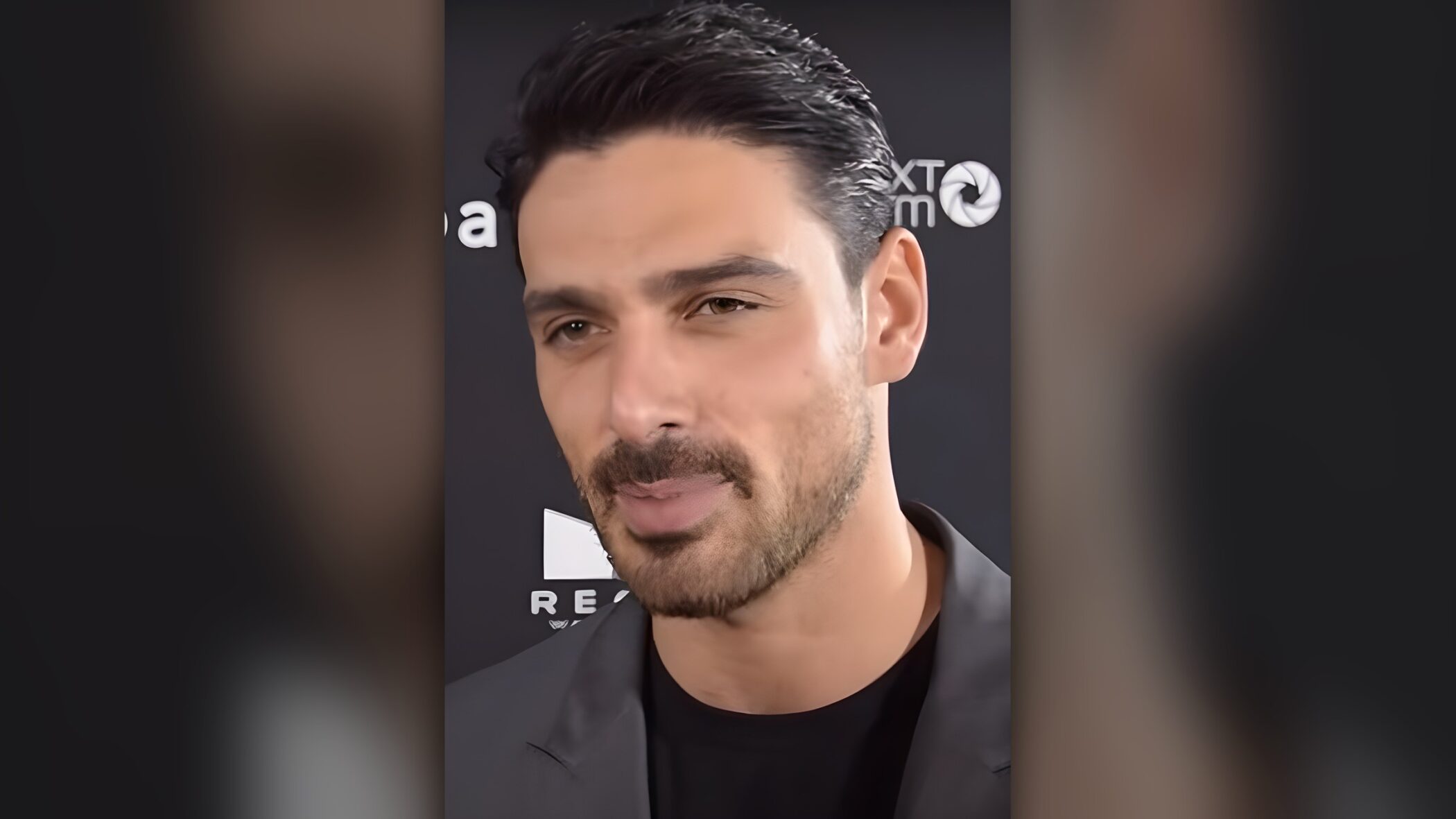 Michele Morrone speaking to the camera at a press event in 2019, in a dark jacket against a logo backdrop