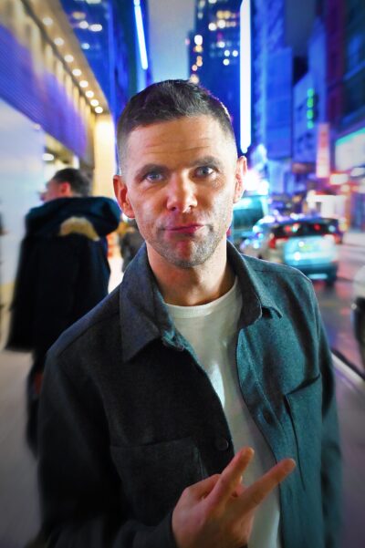 Mikey Day on a New York street at night in 2024, wearing a dark green overshirt and flashing a peace sign