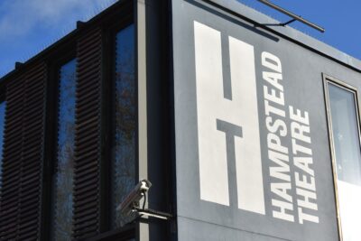 The Hampstead Theatre logo painted on the grey facade of the Swiss Cottage building, with a CCTV camera on the corner