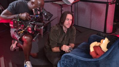Voice actor Matt Mercer seated beside a puppet on a dimly lit set while a camera operator films