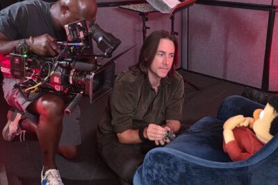Voice actor Matt Mercer seated beside a puppet on a dimly lit set while a camera operator films