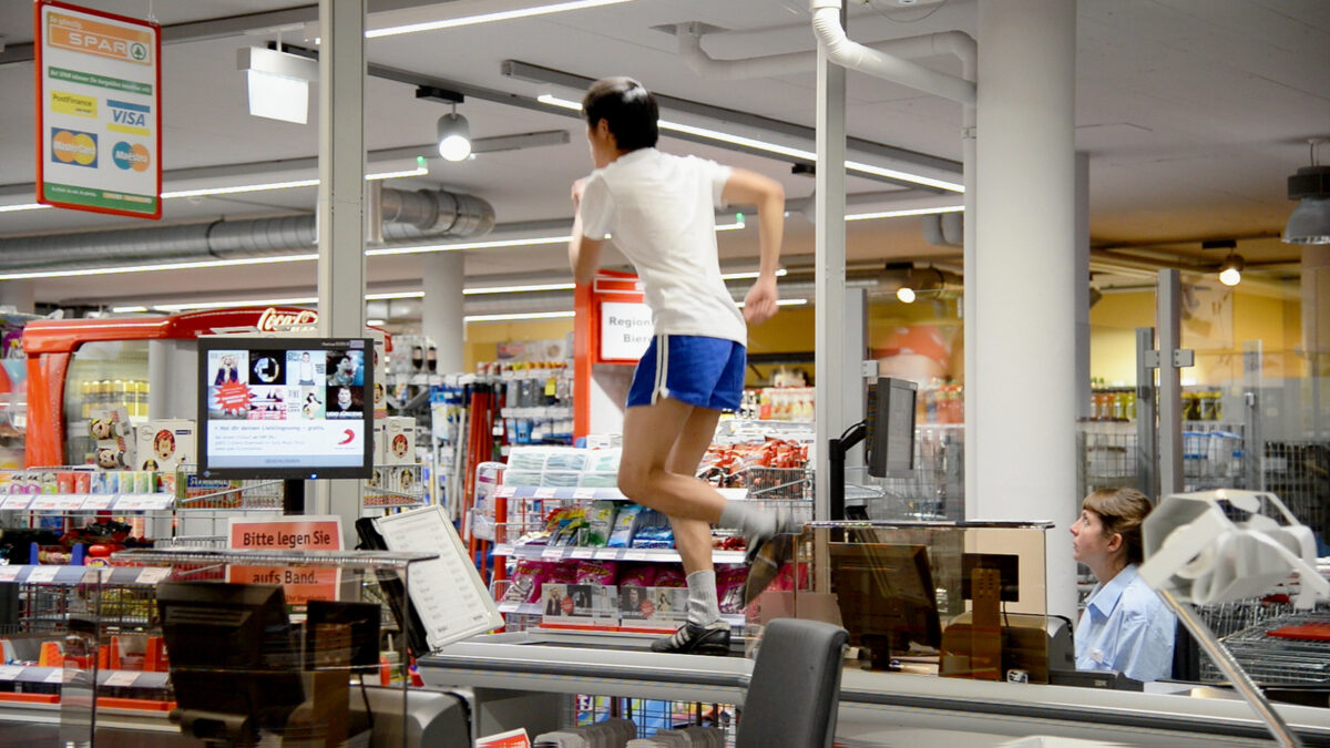 Video still of a performer in white t-shirt and blue shorts running on a supermarket checkout counter as a cashier watches