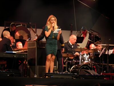 Nicolette van Dam presenting on stage with a microphone in front of a navy band at Sail Amsterdam, 2010