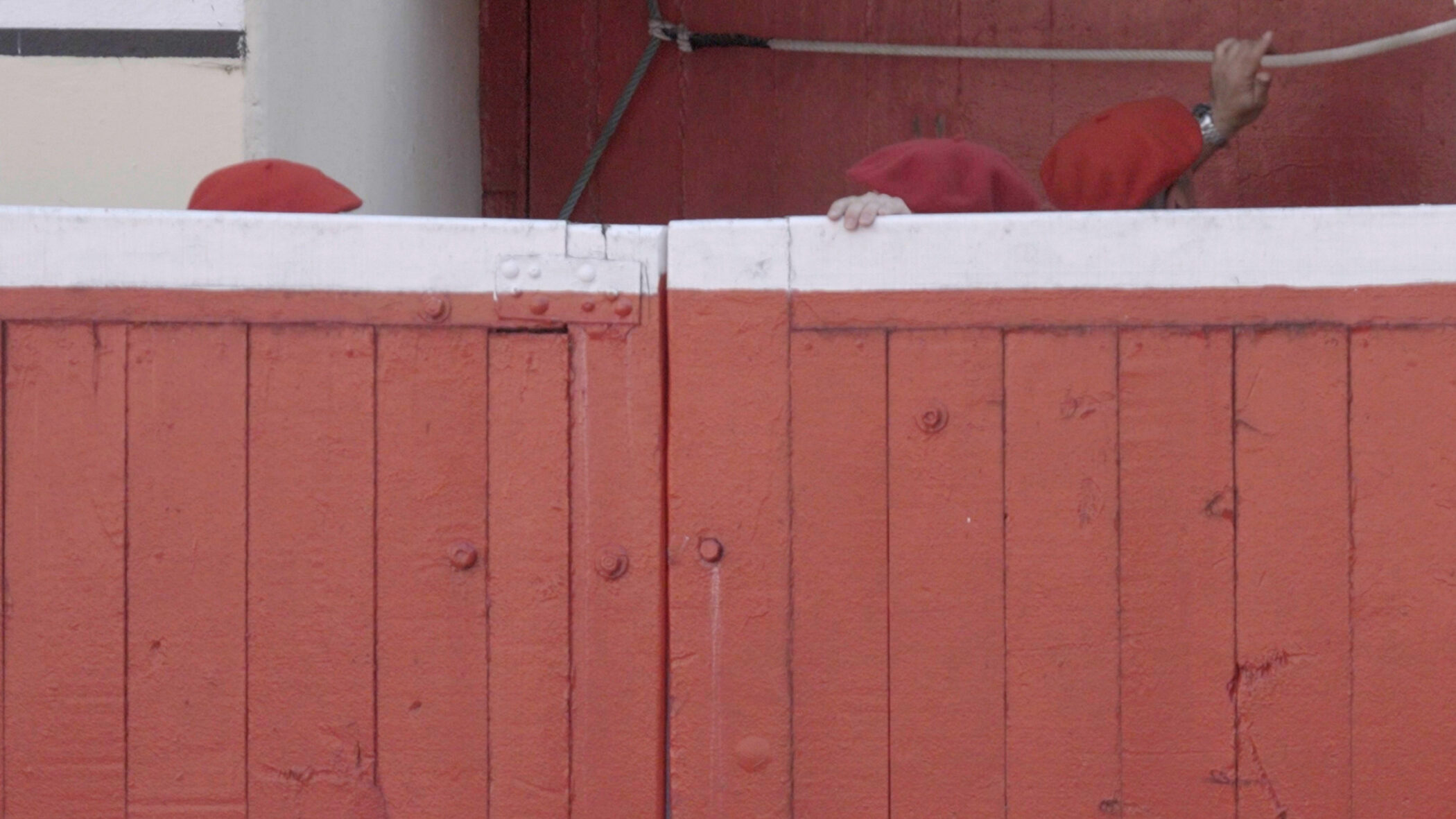 Video still from Paul Pfeiffer Pasodoble: red berets and a hand gripping the top of a red and white bullring barrier