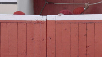 Video still from Paul Pfeiffer Pasodoble: red berets and a hand gripping the top of a red and white bullring barrier