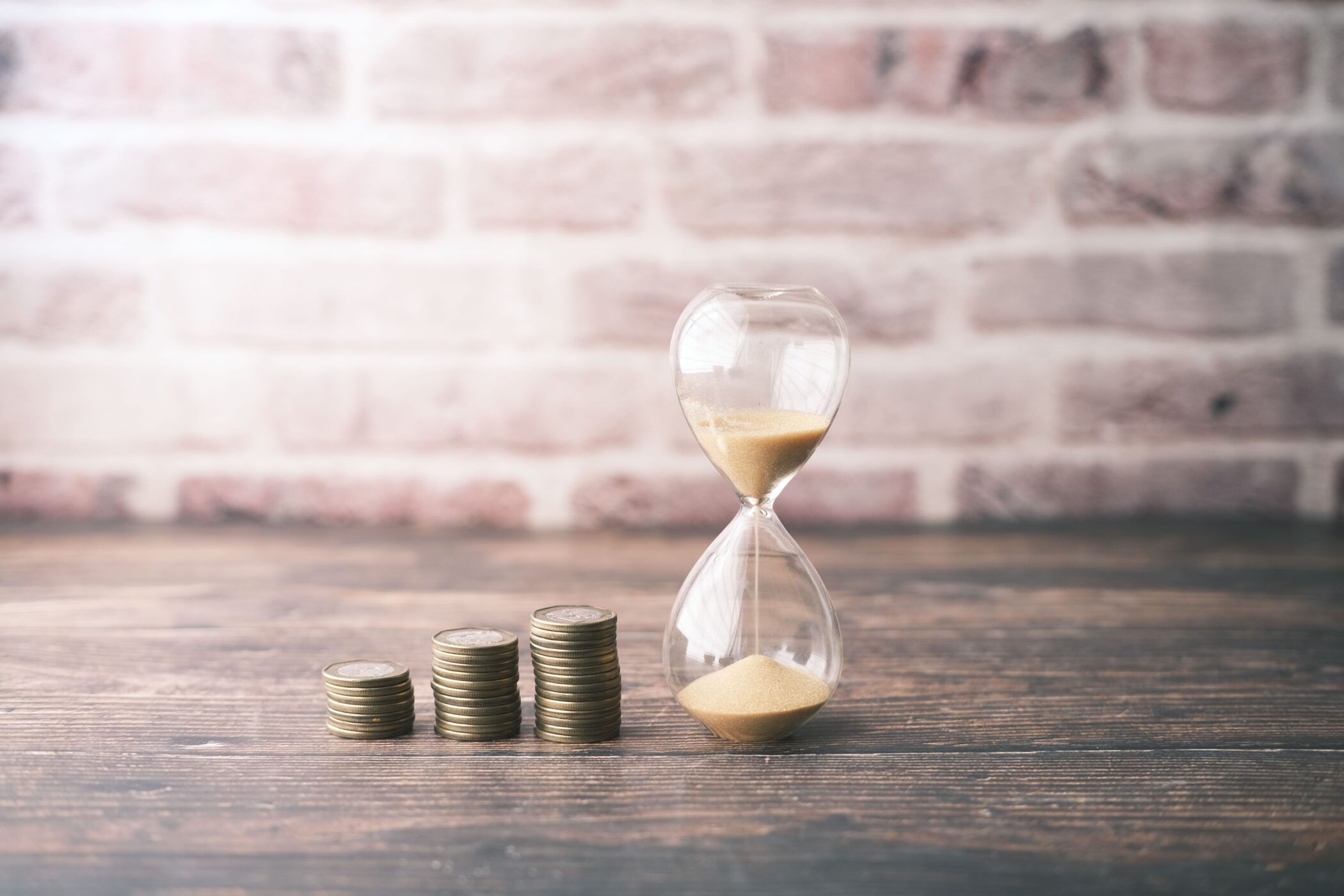 An hourglass with running sand beside three stacks of coins growing in height on a wooden table