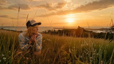 Sophie Woods in a cap, resting her chin on her hands in tall grass above the sea at sunset