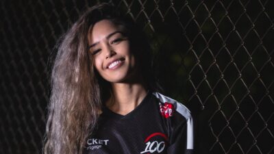 Streamer Valkyrae in a black 100 Thieves jersey in front of a chain-link fence at night