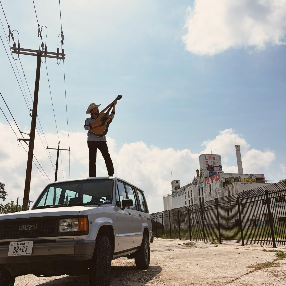 Jerid Reed Morris playing guitar on the roof of a white Isuzu with a BB BS Texas plate