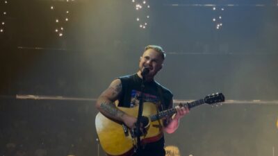 Zach Bryan smiling at the microphone with an acoustic guitar on stage