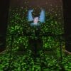 Tröll (photo credit: Tabitha Arthur) Image description: In a pitch black room, a young white man wearing a cap is visible from the shoulders up, illuminated by a square blue light shining up onto his face, with his enlarged silhouette visible behind him. Glitchy green lights are superimposed across the entire frame, and in the top left corner are two pixellated heart icons and several blue vertical bars, as if they were framing a vintage video game.